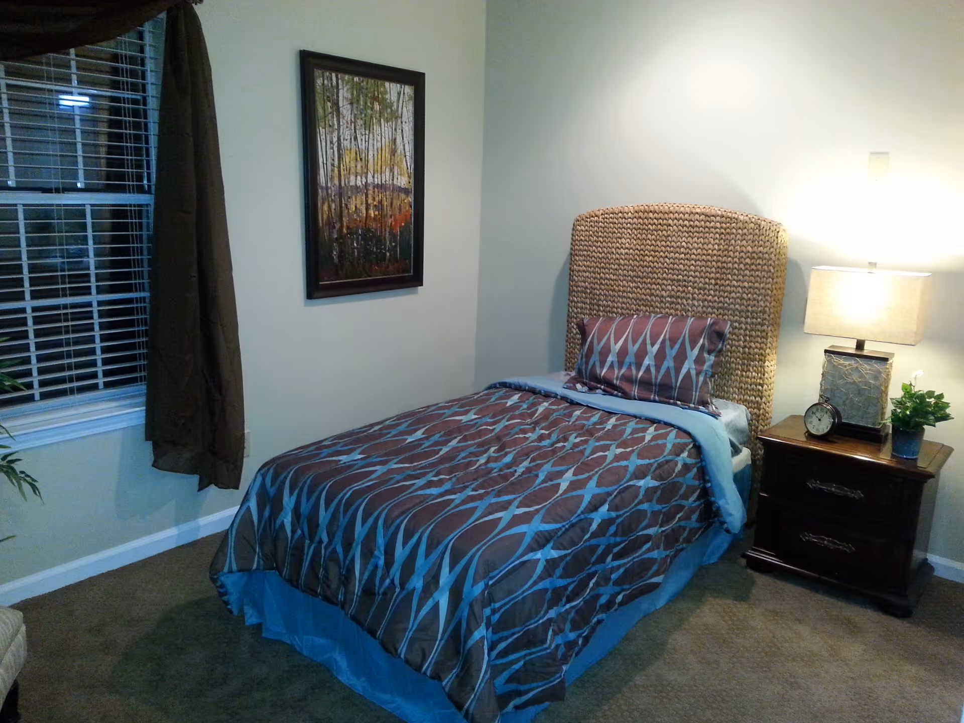 A neatly made single bed with patterned bedding, a woven headboard, and a bedside table with a lamp and clock in a small bedroom.