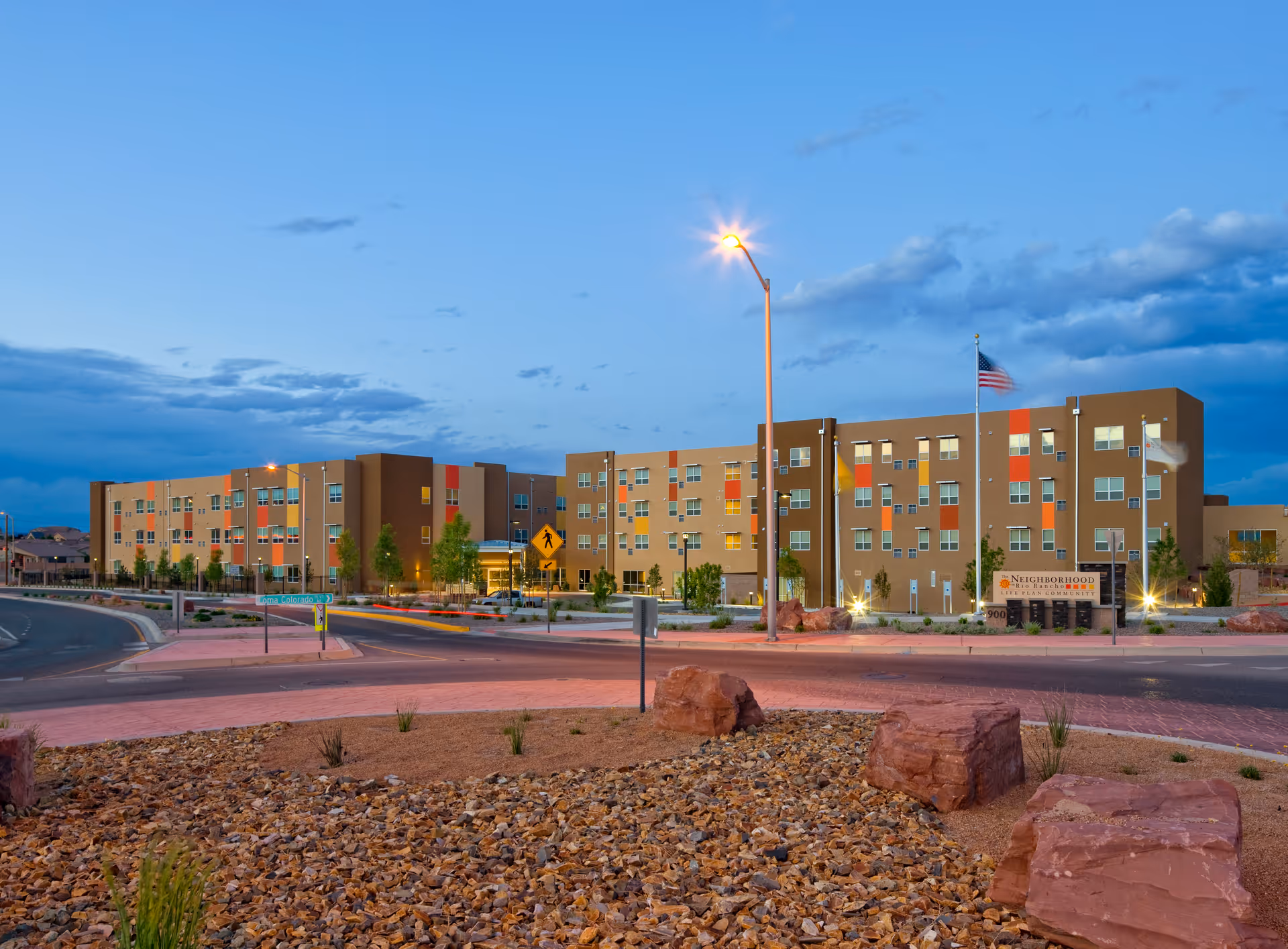 Exterior front view of a modern multi-story senior living community building at dusk with landscaped roundabout and an American flag.
