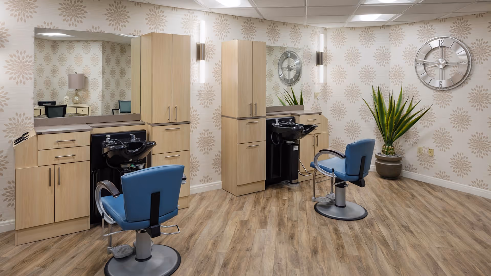 Interior of a salon area with two blue salon chairs in front of black hair washing sinks. The room has light wood cabinetry, large mirrors, patterned wallpaper, a large wall clock, and a potted plant in the corner. The floor is wood-style laminate.