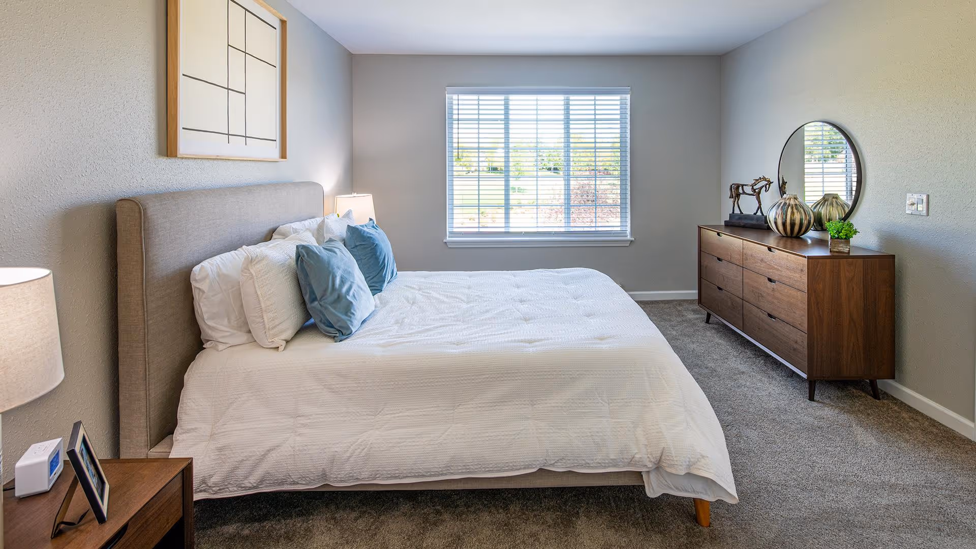 A bright and clean bedroom with a large bed featuring white bedding and blue pillows. There is a wooden nightstand with a lamp and a small digital clock on the left side of the bed. On the right side of the room, there is a wooden dresser with decorative items including a round mirror, a horse figurine, and a small plant. A large window with blinds lets in natural light, illuminating the neutral-colored walls and carpeted floor.