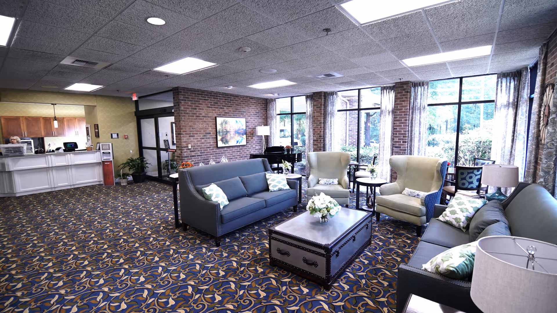Spacious lobby/lounge with sofas, armchairs, a coffee table, a reception desk, and large windows overlooking greenery.