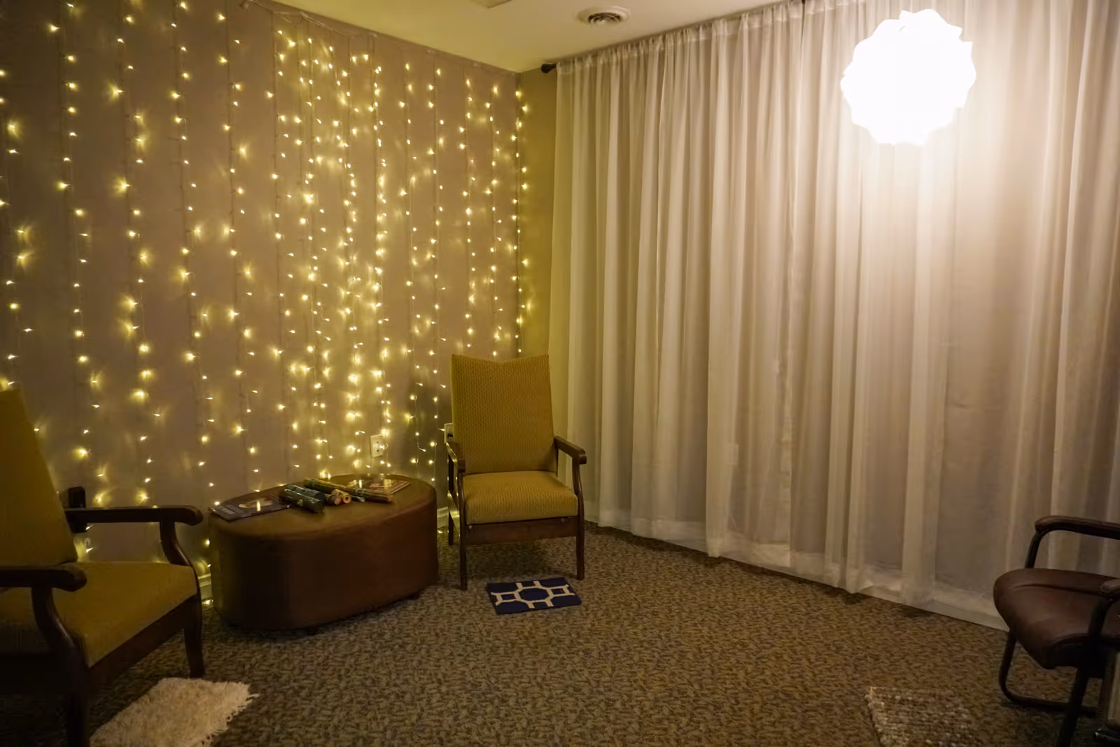 A cozy room with three chairs arranged around a small oval table. The wall behind one chair is decorated with hanging string lights, and the opposite wall has white curtains. A bright, round ceiling light hangs above, illuminating the space softly.