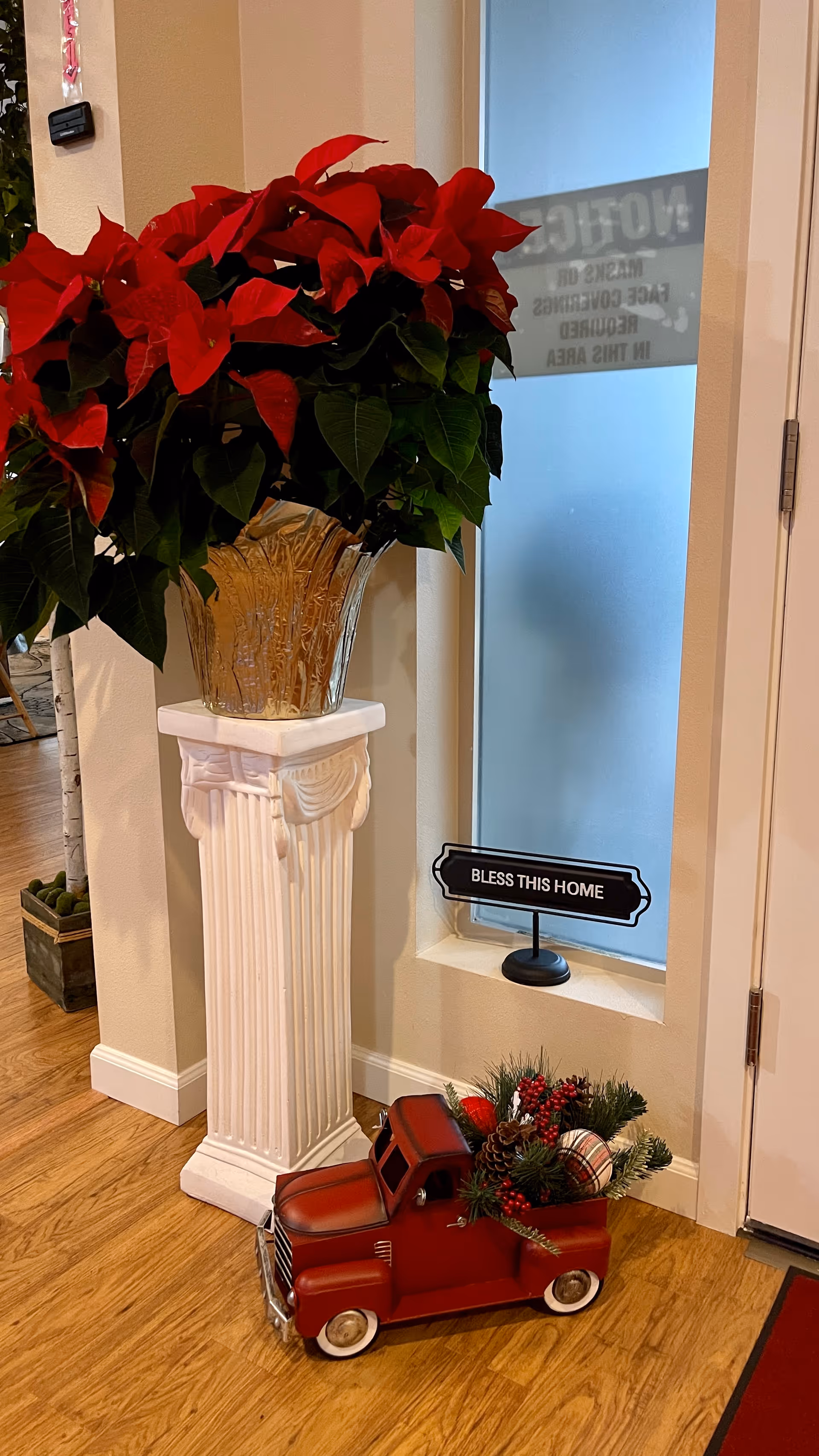 A large red poinsettia on a white pedestal beside a frosted window with a 'Bless This Home' sign and a small red decorative truck filled with greenery on the floor.