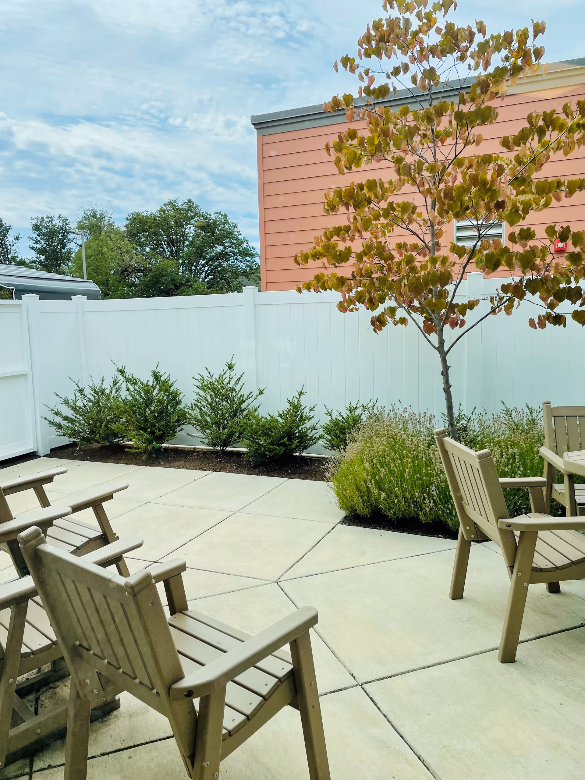 Outdoor patio area with beige wooden chairs arranged around a concrete paved space. There is a small tree with autumn-colored leaves and some green shrubs along a white fence. A building with orange siding is visible in the background under a partly cloudy sky.