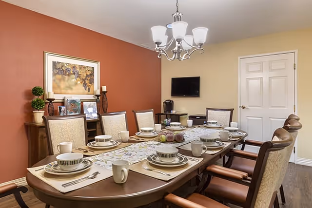 Dining room with a long wooden table set for eight people with plates, bowls, cups, and utensils. The room has a red accent wall with a framed artwork and decorative plants on a sideboard. A chandelier hangs above the table, and a flat-screen TV is mounted on a beige wall near a closed white door.