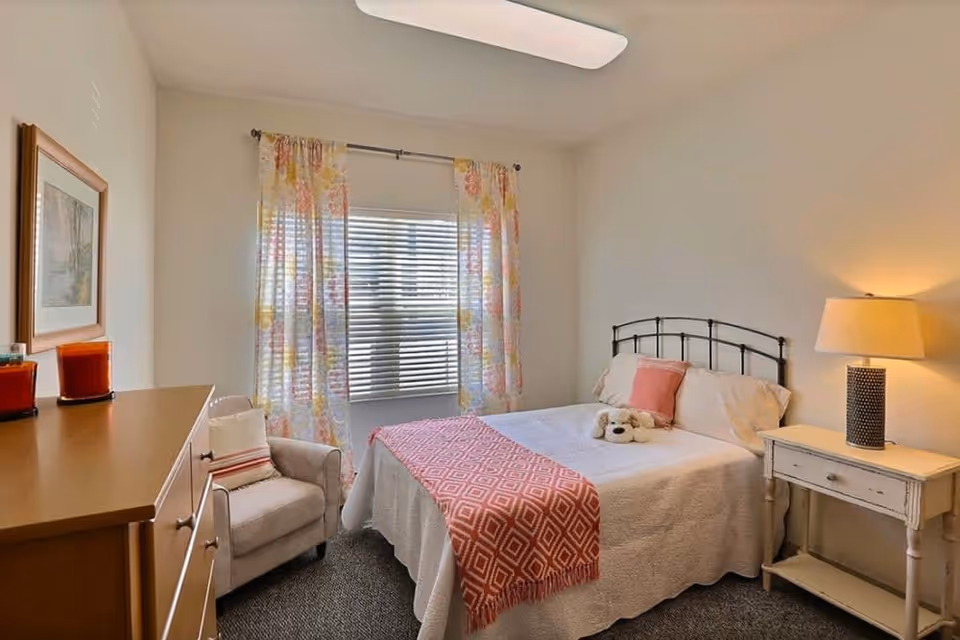 A cozy bedroom with a bed covered in white bedding and a pink patterned throw blanket. There is a small stuffed dog on the bed along with a pink pillow. Next to the bed is a white nightstand with a lamp. A beige armchair with a striped pillow sits next to a wooden dresser with two red candles on it. The window has floral curtains and blinds, and the walls are painted light beige.