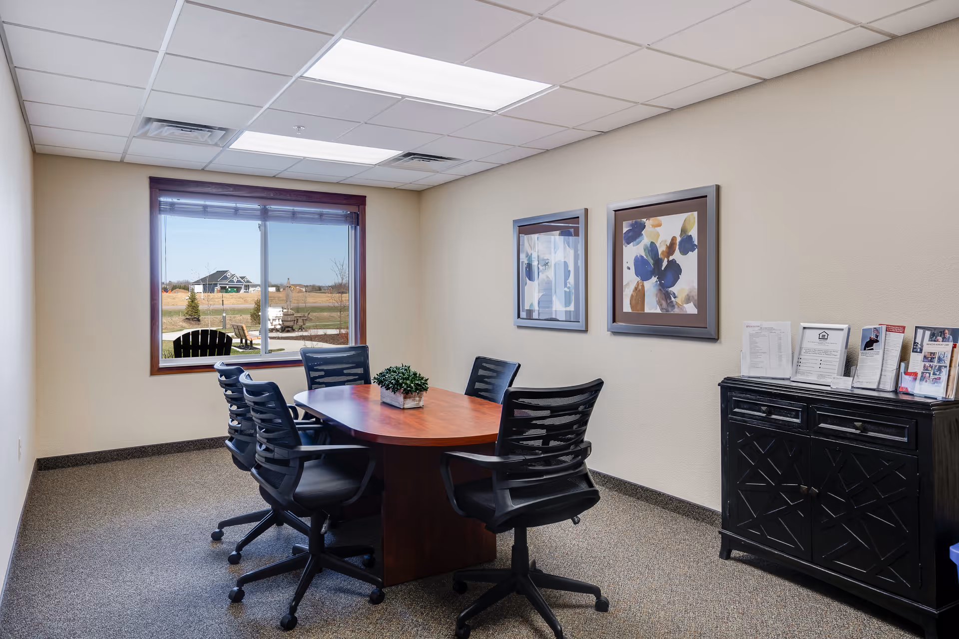 A small conference room with a wooden table surrounded by six black office chairs on wheels. There is a window showing an outdoor view with a chair and some greenery. On the wall are two framed abstract floral paintings. A black cabinet with brochures and papers on top is against the right wall.