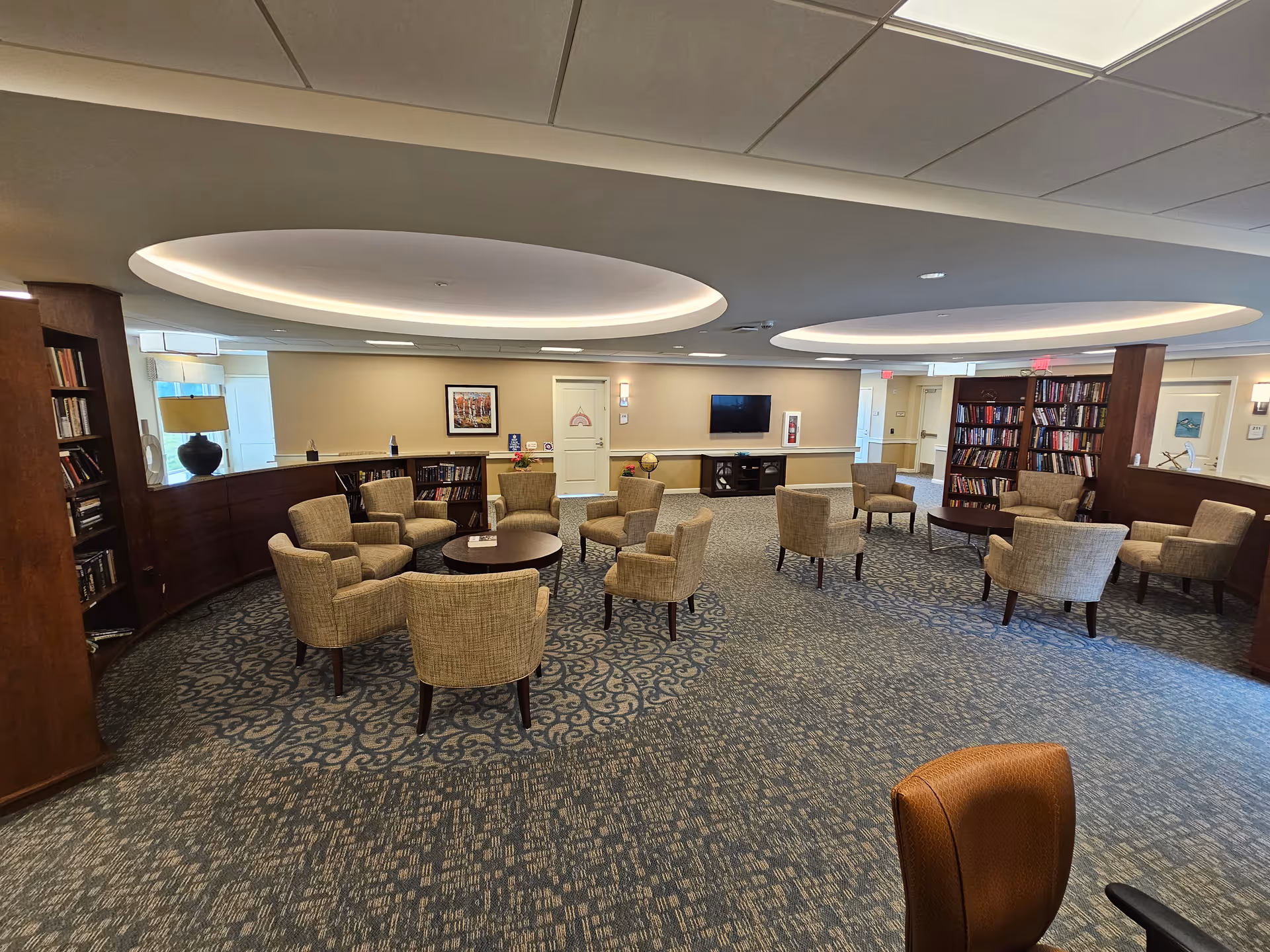 A spacious senior living facility common area with multiple beige upholstered chairs arranged around small round tables. The room features patterned carpet, built-in wooden bookshelves filled with books, a wall-mounted TV, and soft ceiling lighting with circular recessed fixtures. The walls are light-colored with framed artwork and a door in the center background.