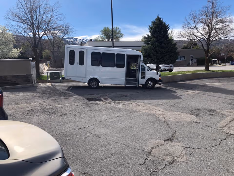 A white passenger shuttle parked in a paved lot in front of a low building with trees and mountains in the background.