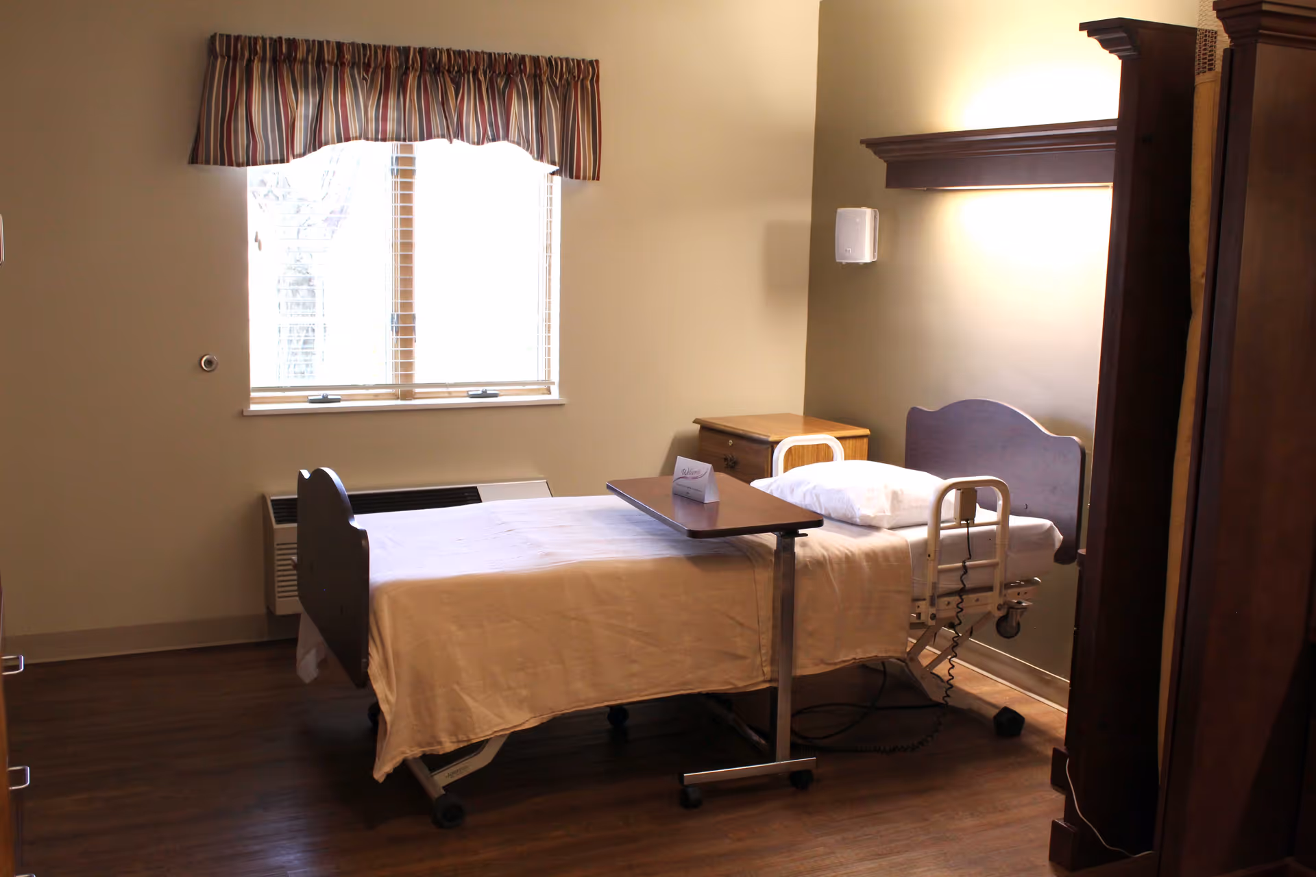 A simple senior living bedroom with a single hospital-style bed covered with a beige blanket and a white pillow. There is a small overbed table on wheels positioned over the bed. The room has wooden flooring, a window with striped valance curtains, a wooden nightstand, and a wooden wardrobe. The walls are painted a light beige color.