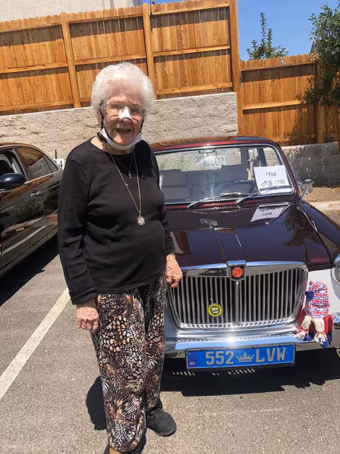 An elderly woman with white hair, glasses, and a black top stands smiling next to a vintage maroon MG car parked in an outdoor parking lot with a wooden fence in the background. The car has a blue license plate and a sign on the windshield indicating it is a 1966 MG 1100.