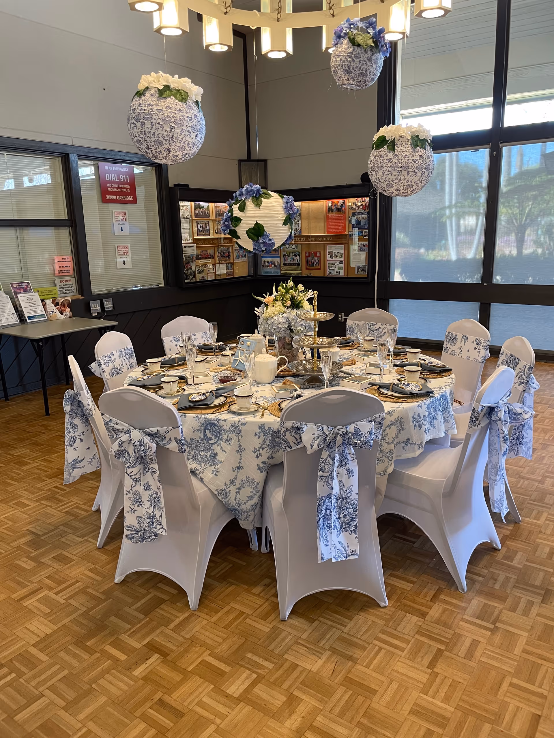 Round dining table set for a formal meal with white and blue floral tablecloth and chair covers, floral centerpiece and hanging decorative spheres in a community dining room.