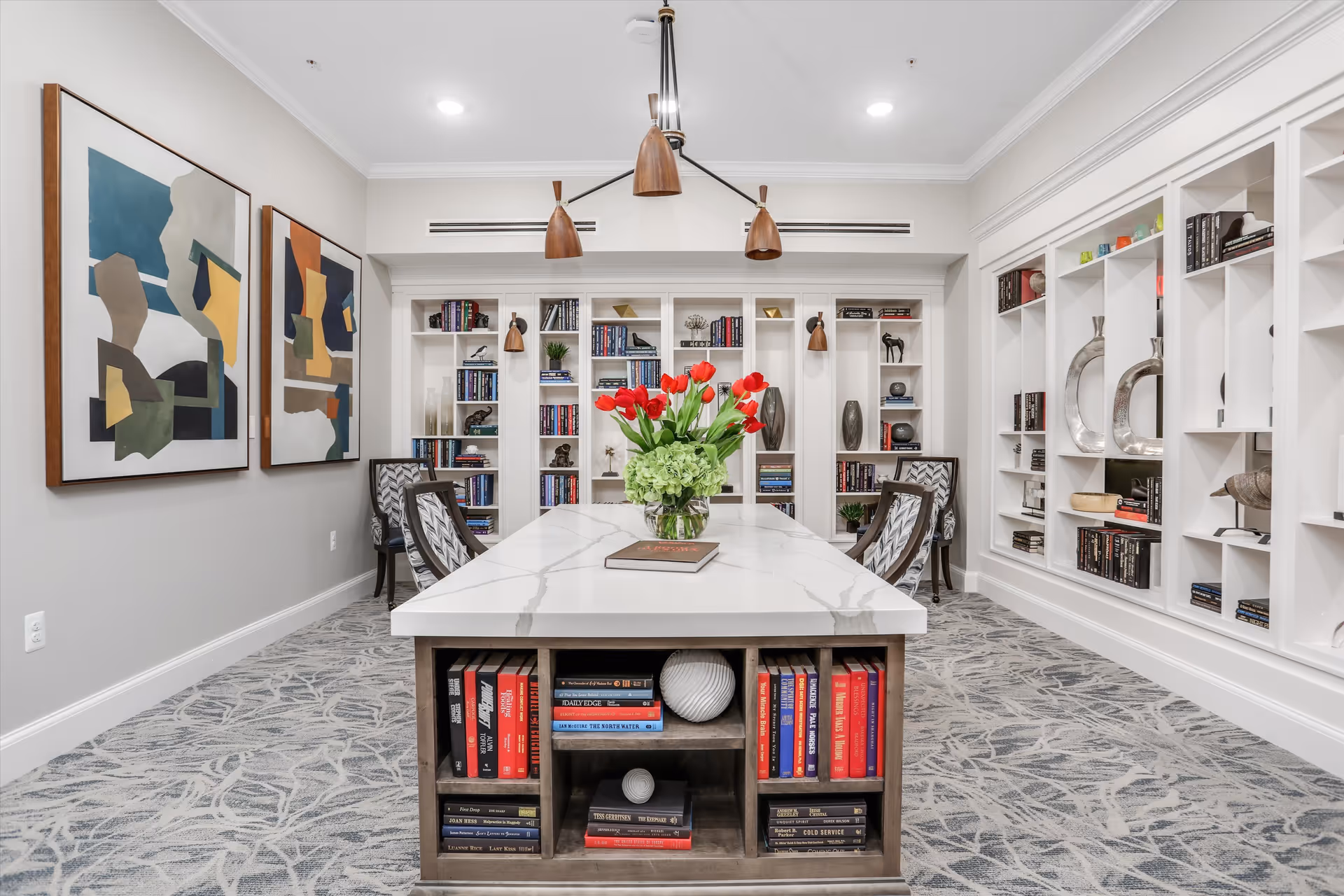 A modern and elegant library or reading room with a large white marble table in the center, surrounded by patterned chairs. The table has built-in shelves filled with books and decorative items. The walls feature built-in white bookshelves filled with books and decorative objects, and two abstract paintings hang on the left wall. A contemporary chandelier with wooden shades hangs above the table, and a vase with red tulips and green hydrangeas is placed on the table.