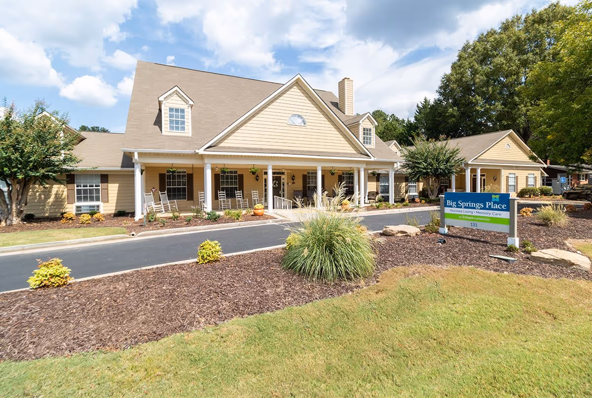Exterior view of a single-story assisted living facility with beige siding, a gabled roof, and a covered porch with rocking chairs. There are landscaped bushes and trees around the building, and a sign in front reads 'Big Springs Place Assisted Living Memory Care'. The sky is partly cloudy.