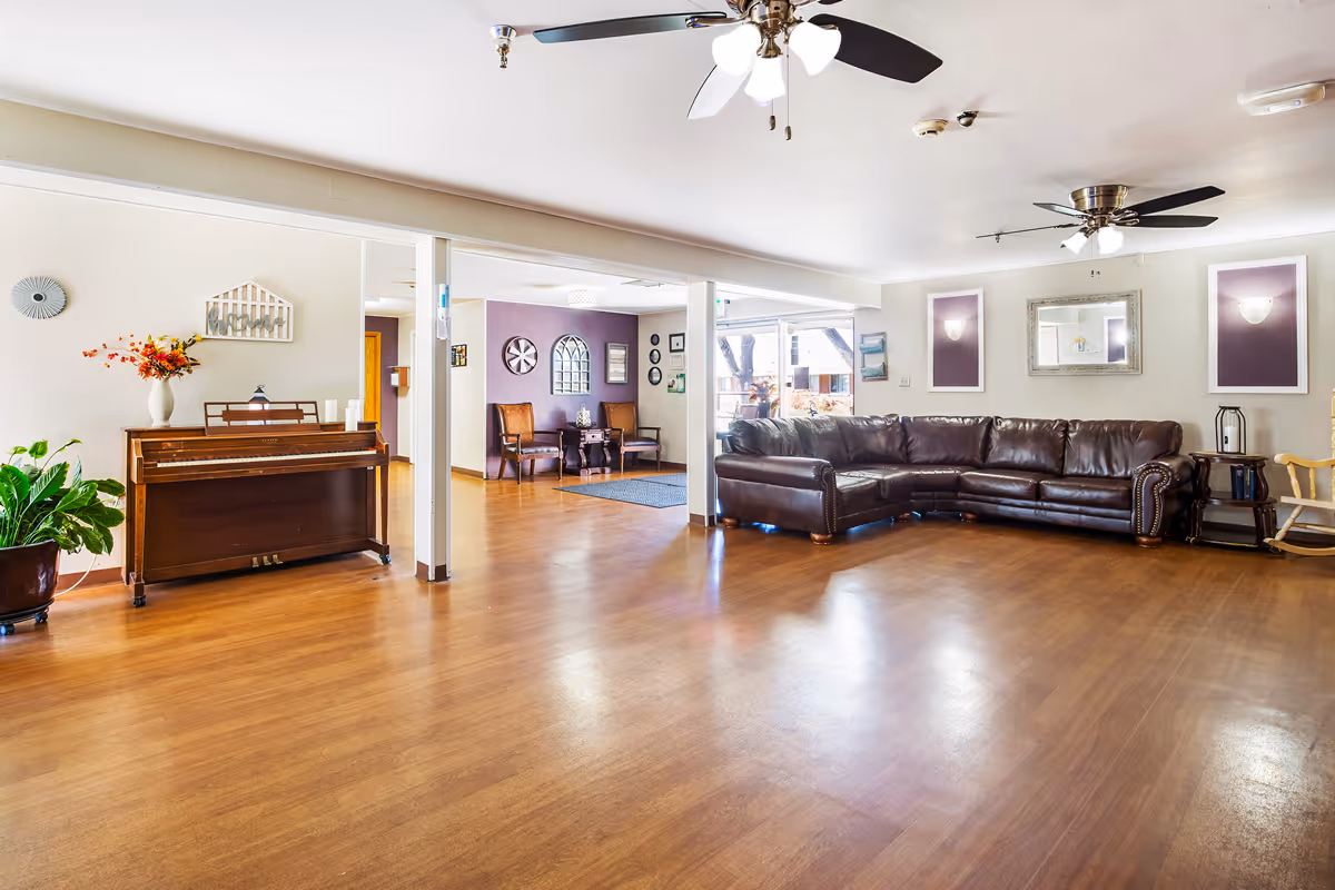 A spacious, well-lit living room area with wooden flooring, a large dark brown leather sectional sofa, two ceiling fans with lights, a wooden piano with a vase of flowers on top, and several chairs and decorative wall art. There are large windows letting in natural light and a small rocking chair near the sofa.