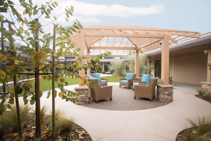 Outdoor seating area with four wicker chairs featuring blue patterned cushions arranged in a circle under a wooden pergola. The area is surrounded by a concrete pathway, small trees, and green grass within a courtyard setting.
