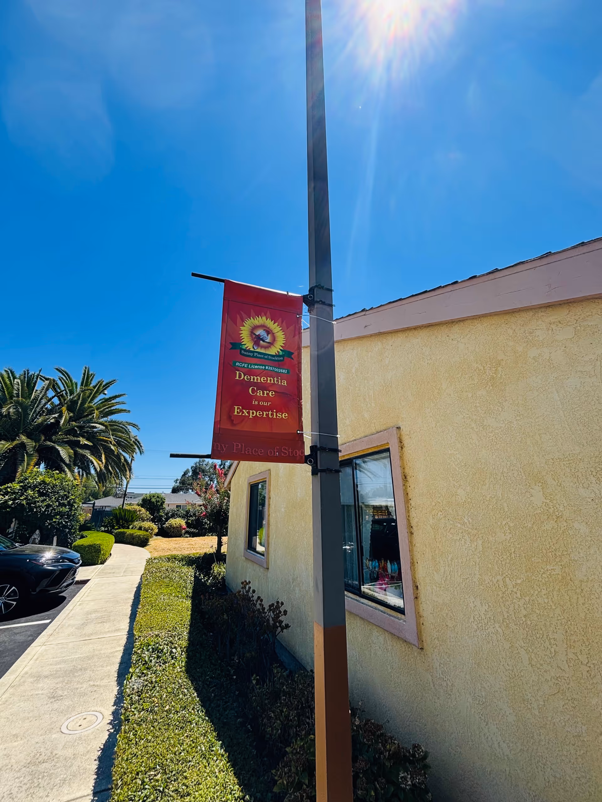 Exterior of a yellow senior living building with a sidewalk, landscaping, and a pole banner that reads 'Dementia Care is our Expertise'.