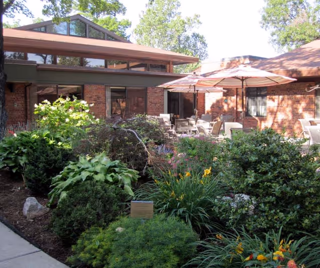 Outdoor patio area at The Mary Culver Home for Visually Impaired Women featuring a garden with various green plants and flowers in the foreground, and brick buildings with large windows and patio umbrellas with chairs in the background.