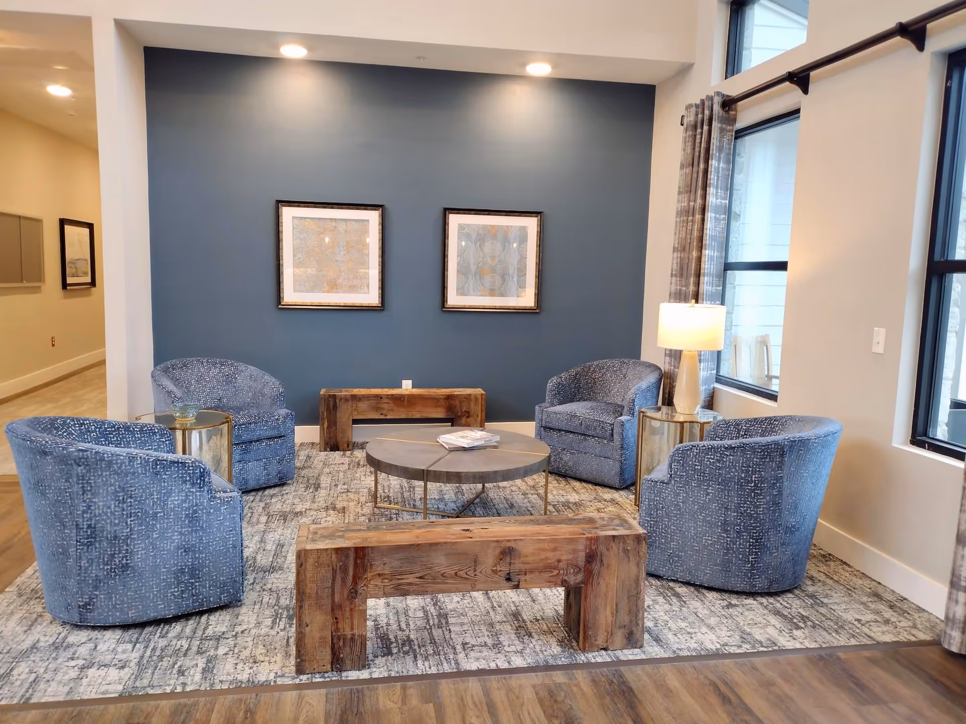 A cozy sitting area with four blue upholstered armchairs arranged around a round coffee table on a patterned rug. There are two wooden benches, one in the foreground and one against a blue accent wall with two framed artworks. A table lamp on a small round side table is placed near a window with curtains.