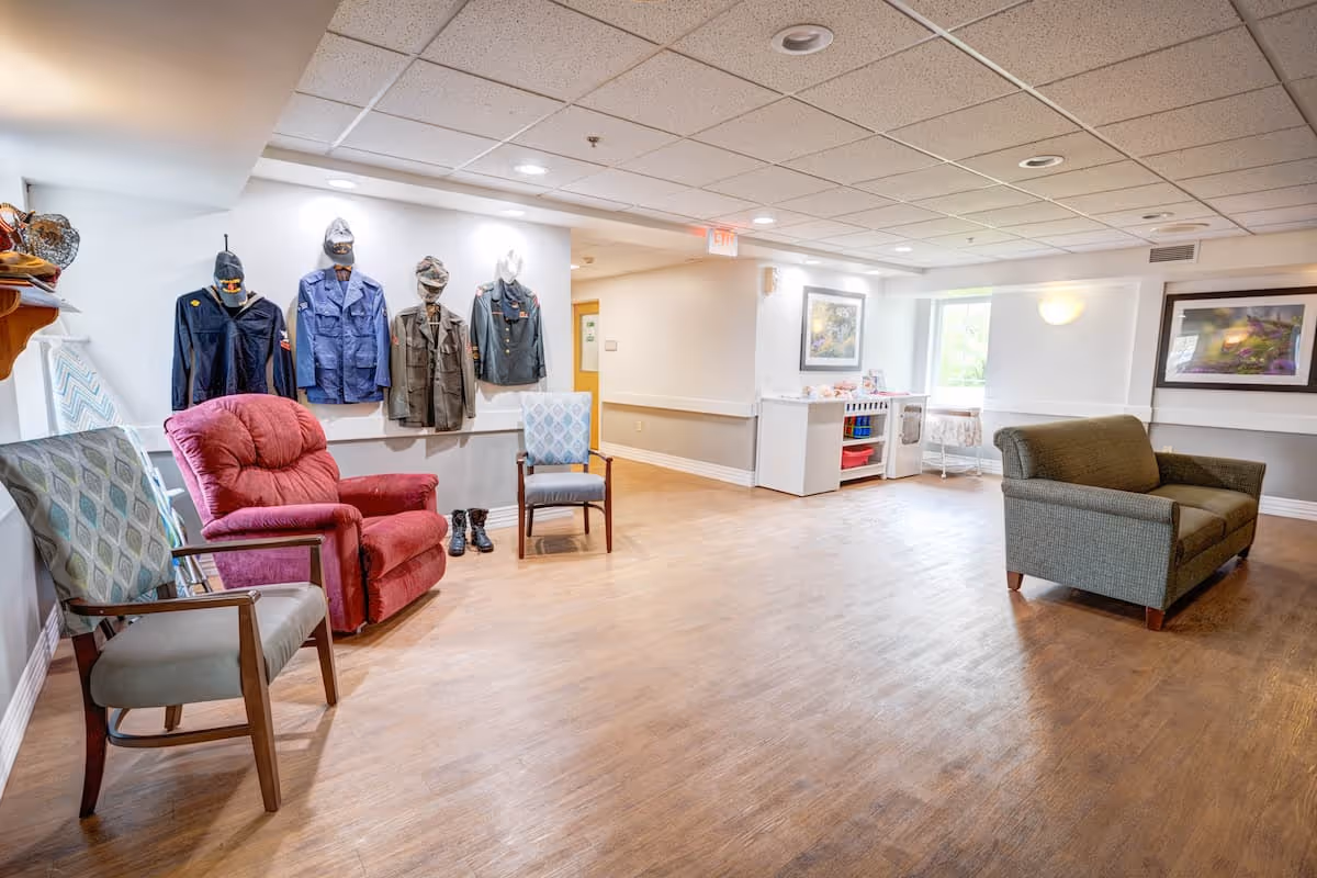 A spacious and well-lit common area in a senior living facility with wooden flooring, several chairs including a red recliner and two patterned armchairs, a green loveseat, and a display of military uniforms and boots mounted on the wall. The room has white walls, ceiling lights, framed artwork, and a window letting in natural light.