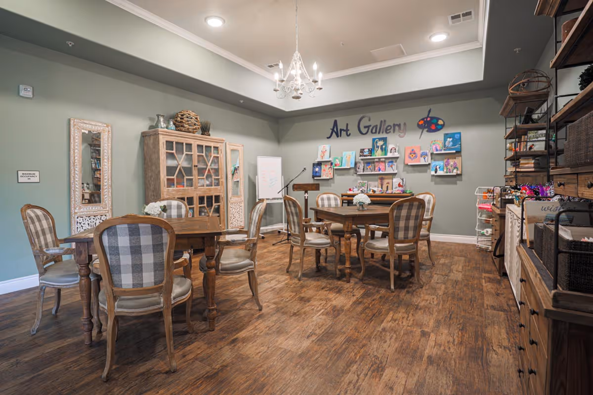 A cozy art gallery room in an assisted living facility with wooden tables and chairs featuring plaid upholstery. The walls are decorated with colorful artwork and a sign that reads 'Art Gallery'. There is a wooden cabinet, a whiteboard, and shelves with various supplies. The room has wooden flooring and a chandelier hanging from the ceiling.