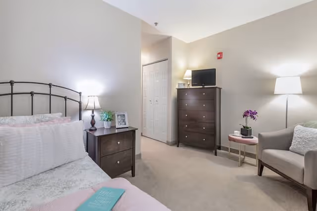 A cozy bedroom with a bed on the left side featuring white and pink bedding. Next to the bed is a dark wooden nightstand with a lamp, a small plant, and a framed photo. Across from the bed is a tall dark wooden dresser with a small TV and a lamp on top. To the right, there is a beige armchair with a white pillow and a small round side table holding a purple orchid and a cup. The room is softly lit with a floor lamp in the corner and has beige walls and carpet.