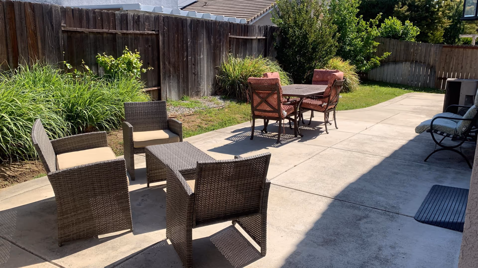 A sunlit backyard patio with wicker seating and a dining table on a concrete slab next to a wooden fence and landscaping.