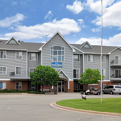 Front exterior of a multi-story senior living facility building with a central gabled entrance, trees, and a circular driveway.
