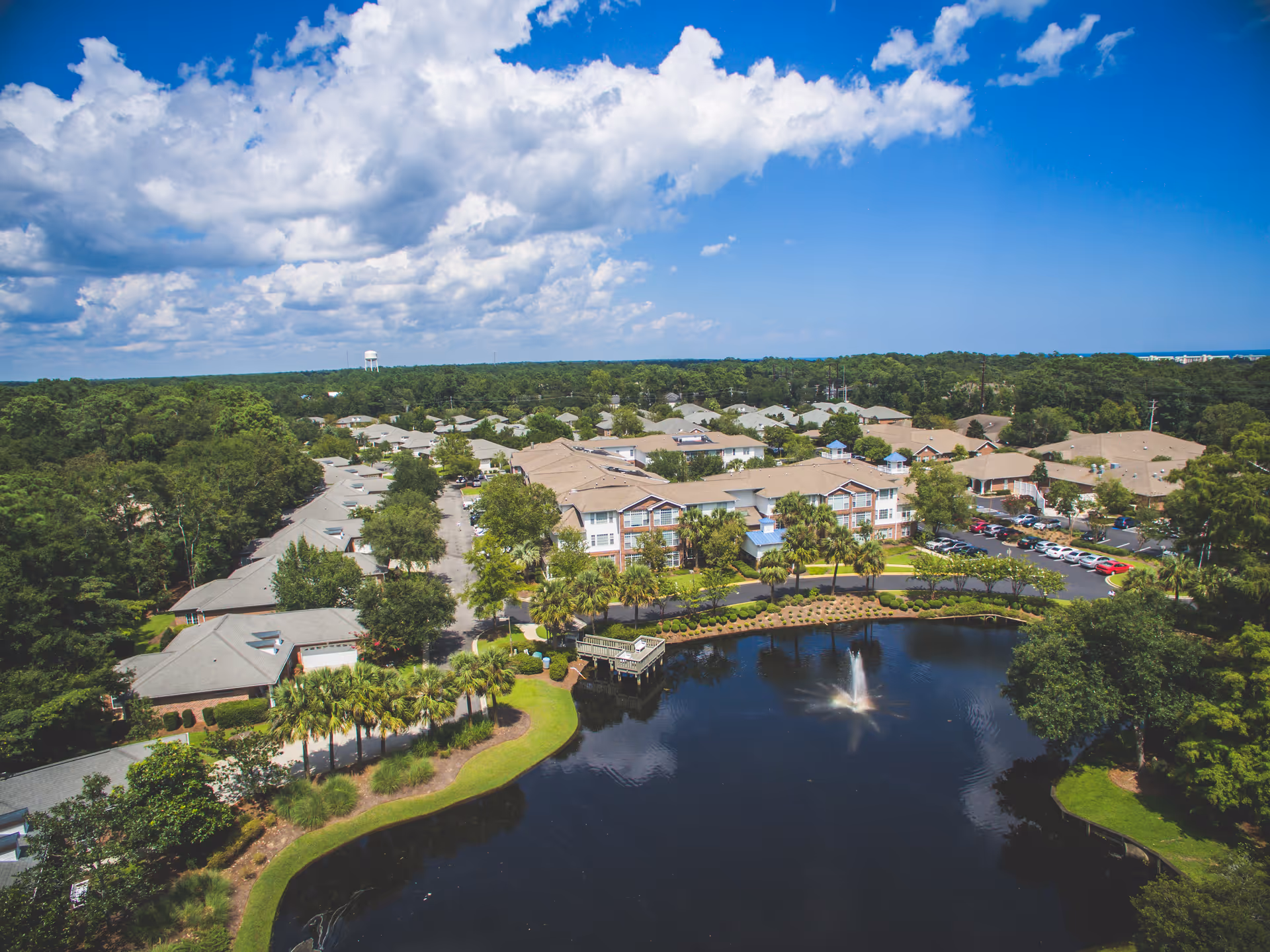 Aerial view of a lakeside senior living campus with multiple buildings, a fountain in the pond, parking, and surrounding trees.