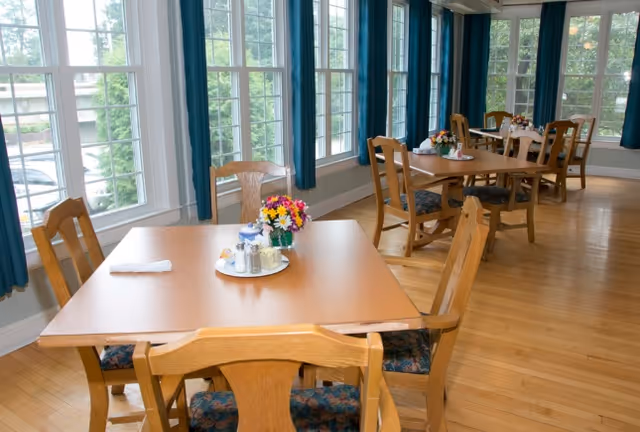 A bright dining room with wooden tables and chairs arranged neatly. Each table has a small centerpiece with colorful flowers and condiments. Large windows with blue curtains allow natural light to fill the room, and the floor is made of polished wood.