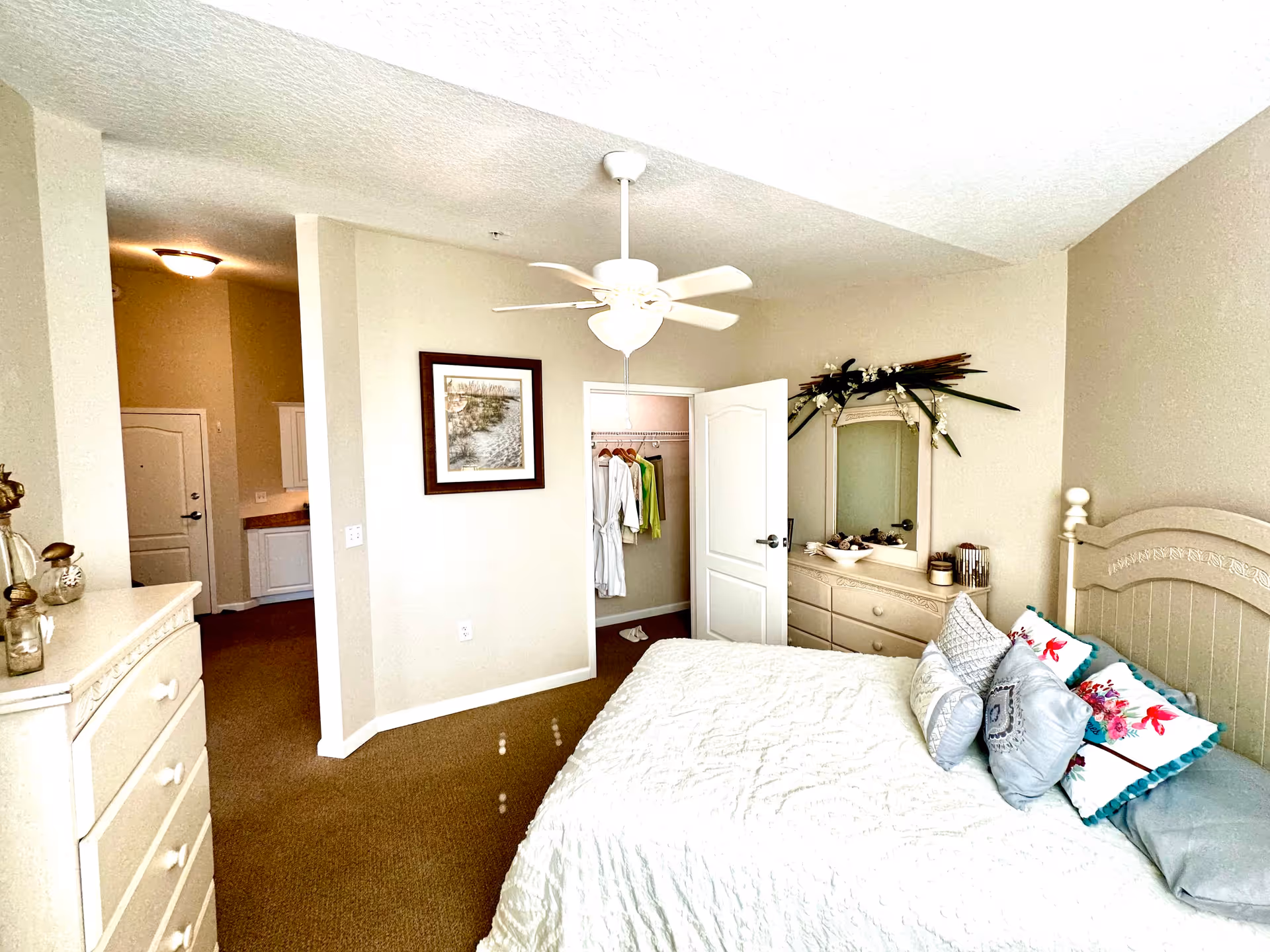 A bright bedroom in a senior living facility featuring a bed with multiple decorative pillows and a white quilted bedspread. The room includes a dresser with a mirror adorned with floral decorations, a ceiling fan with a light, and an open closet door showing hanging clothes. The walls are painted beige, and there is a framed picture on the wall. The floor is carpeted in brown.