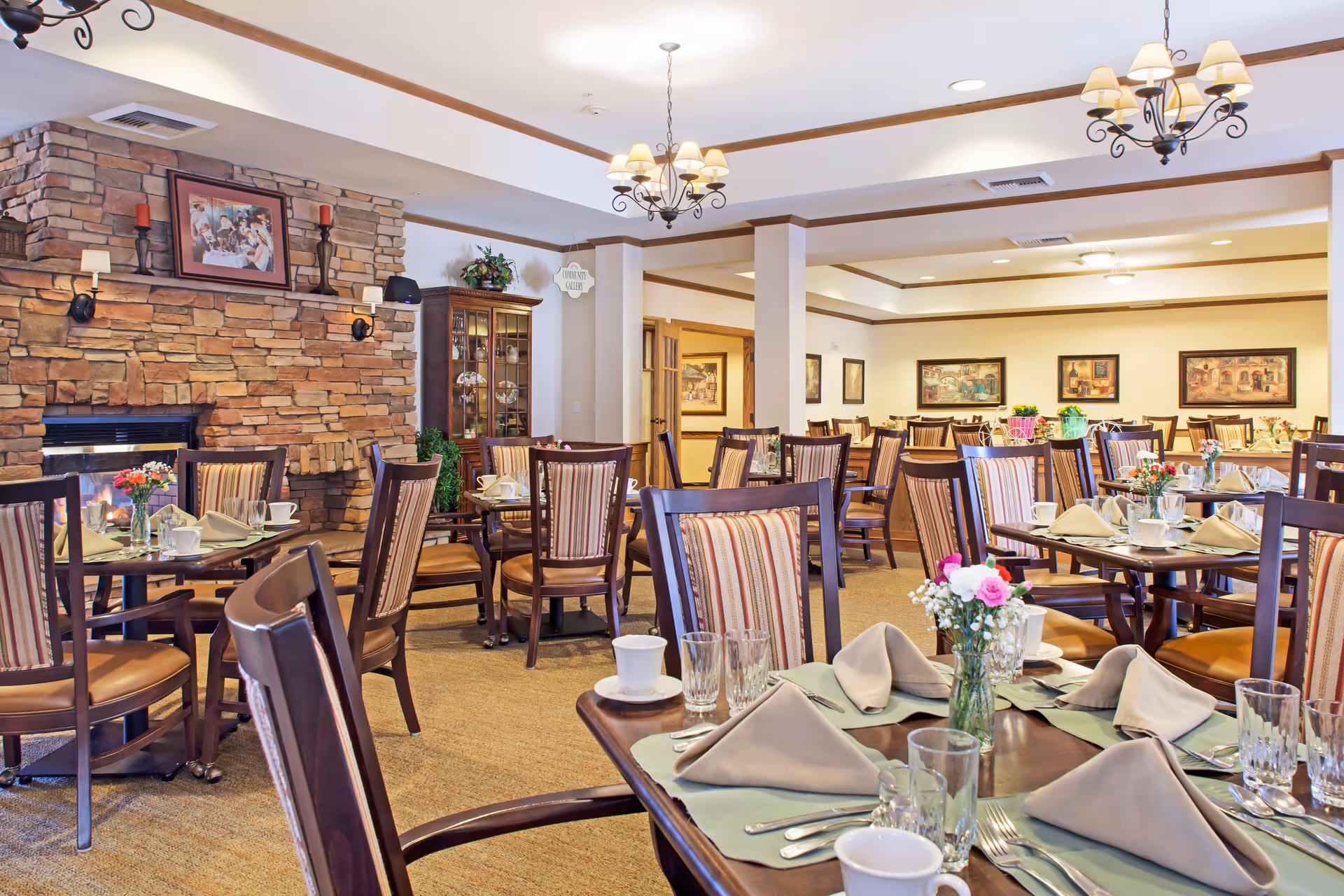A spacious dining room in a senior living facility with multiple wooden tables and chairs arranged neatly. Each table is set with beige napkins, glassware, cups, and silverware. There are small flower arrangements on the tables. The room features a stone fireplace on the left wall, framed artwork, and warm lighting from chandeliers and wall sconces.