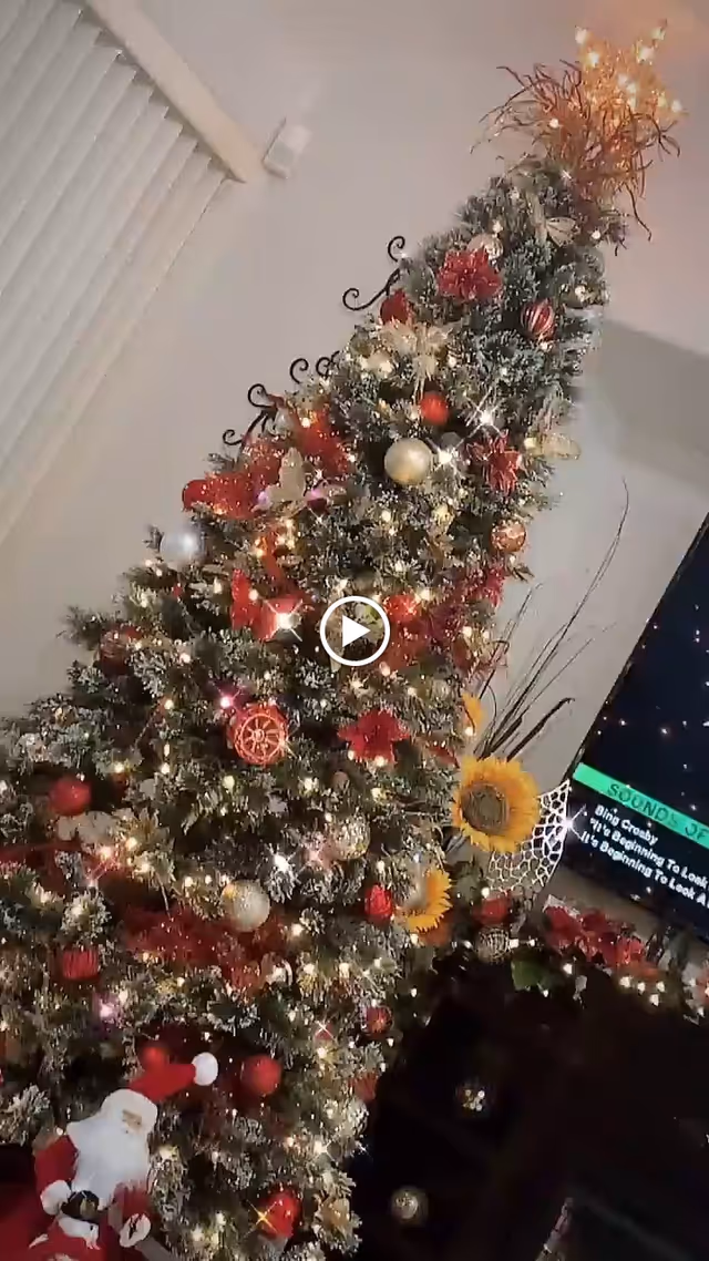 A tall, decorated Christmas tree with lights, red and gold ornaments, and poinsettias stands in a living room corner next to a window with vertical blinds. A Santa Claus figure is placed at the base of the tree. Nearby, a TV screen displays a music playlist, and sunflowers and other decorations are visible on a nearby surface.