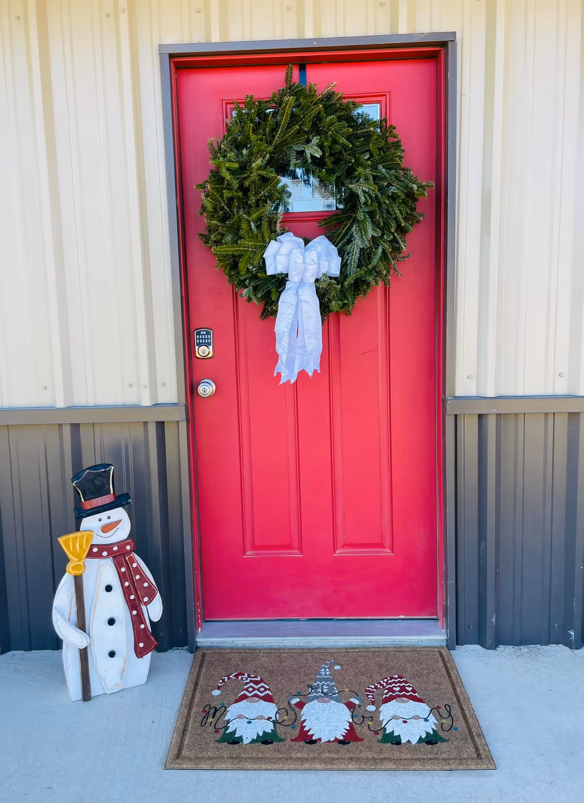 A bright red exterior door decorated with a large green wreath adorned with a white bow. To the left of the door stands a decorative snowman figure holding a broom, wearing a black top hat and a red scarf. In front of the door is a doormat featuring three gnomes wearing festive hats and the word 'Merry' written across it. The door is set in a building with beige and dark gray vertical siding.