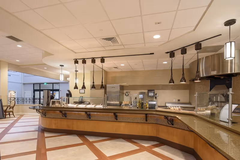 Interior view of a cafeteria-style serving counter with pendant lights, glass sneeze guards, and stainless-steel kitchen equipment.
