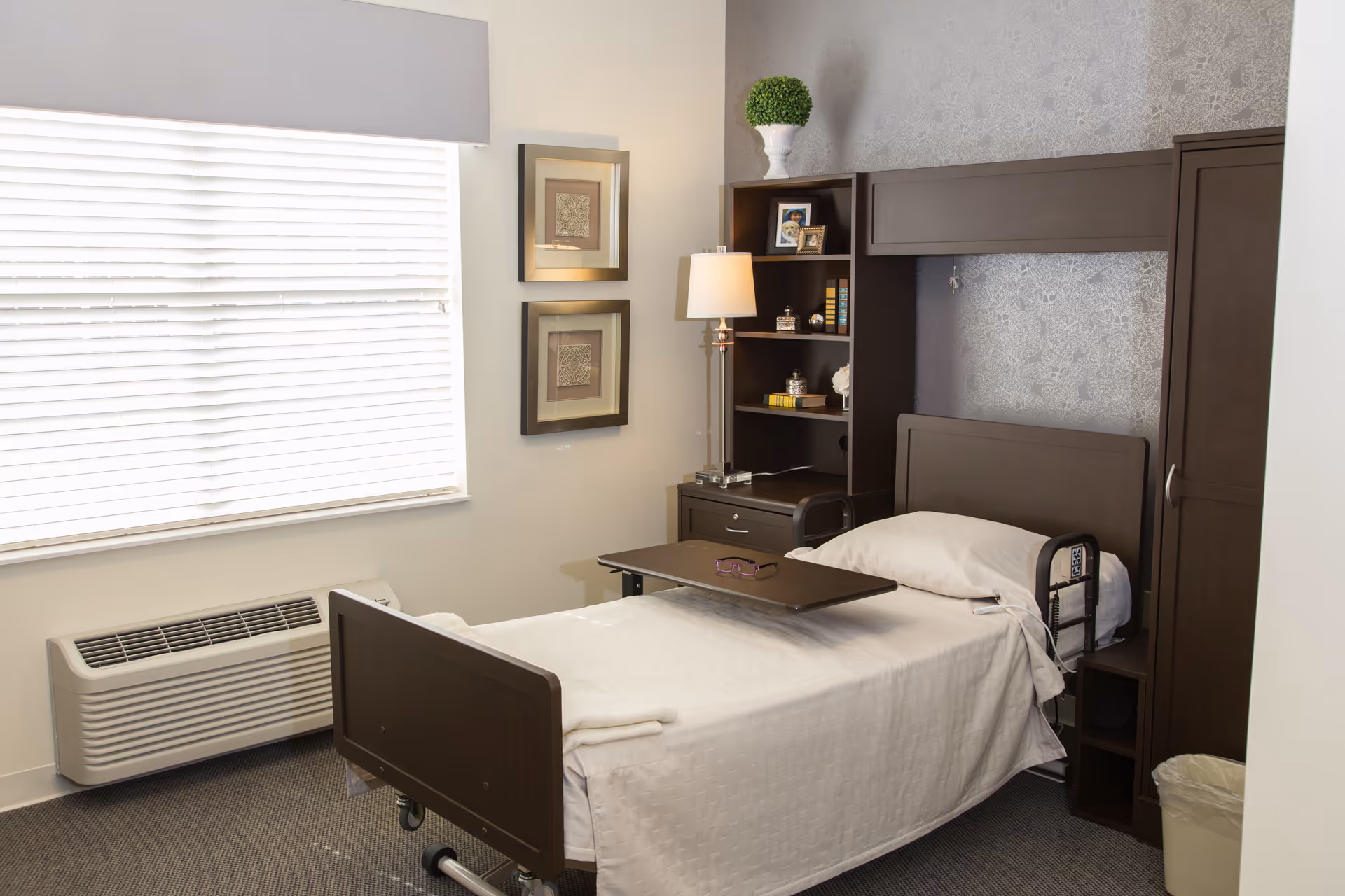 A single bed with white bedding in a senior living facility room. The bed has a dark wooden headboard and footboard, with a small overbed table holding a pair of glasses. Next to the bed is a dark wooden shelving unit with a lamp, books, framed photos, and a decorative plant on top. The room has a window with white blinds, two framed pictures on the wall, a carpeted floor, and a beige wall heater below the window.