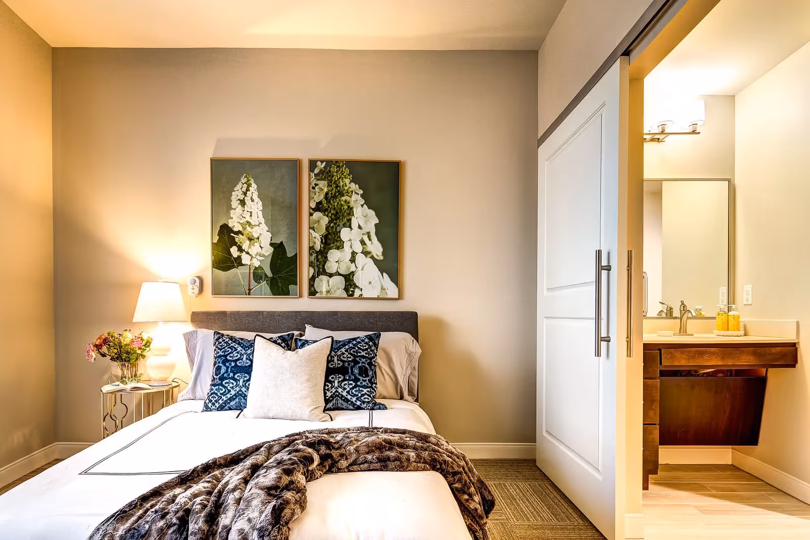 A cozy bedroom with a neatly made bed featuring white and blue pillows and a brown fur throw blanket. There are two framed floral artworks hanging above the bed. A bedside table with a lamp and a vase of flowers is on the left side. To the right, a sliding door opens to a bathroom with a sink, mirror, and light fixture.
