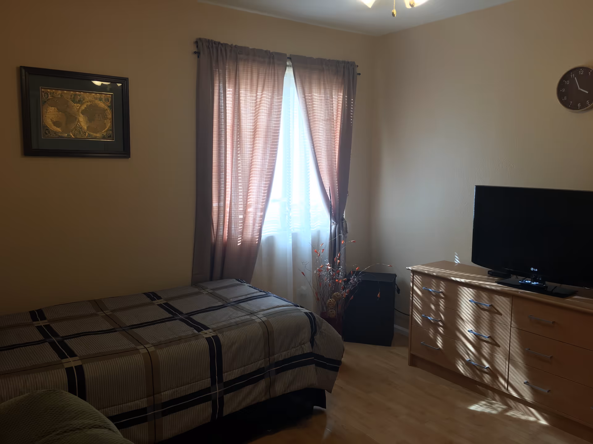 A bedroom with a single bed covered in a plaid bedspread, a window with pink curtains letting in natural light, a wooden dresser with a flat-screen TV on top, a wall clock, and a framed map on the wall.