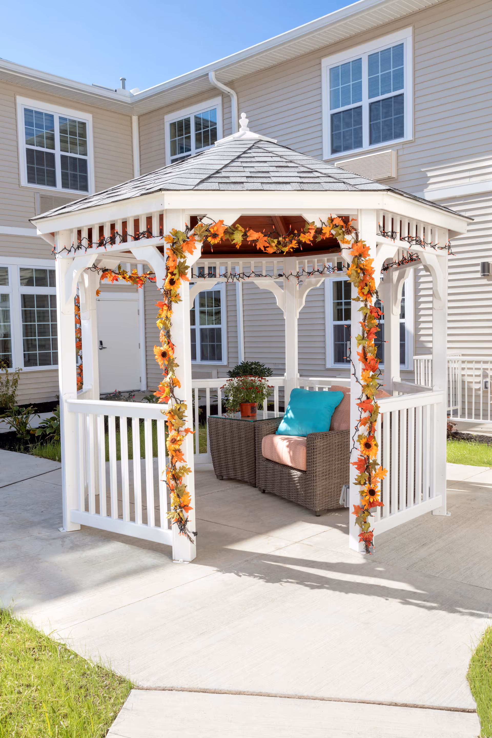 A white wooden gazebo decorated with autumn-themed garlands featuring orange and yellow leaves and sunflowers. Inside the gazebo, there is a wicker chair with a pink cushion and a turquoise pillow, next to a small table with a potted plant. The gazebo is situated on a concrete patio outside a beige building with multiple windows.