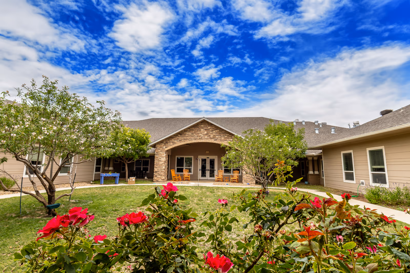 Outdoor courtyard area of Bristol Park at Amarillo Assisted & Living Memory Care featuring a green lawn, blooming red flowers in the foreground, trees, and a building with a covered porch with rocking chairs under a partly cloudy blue sky.