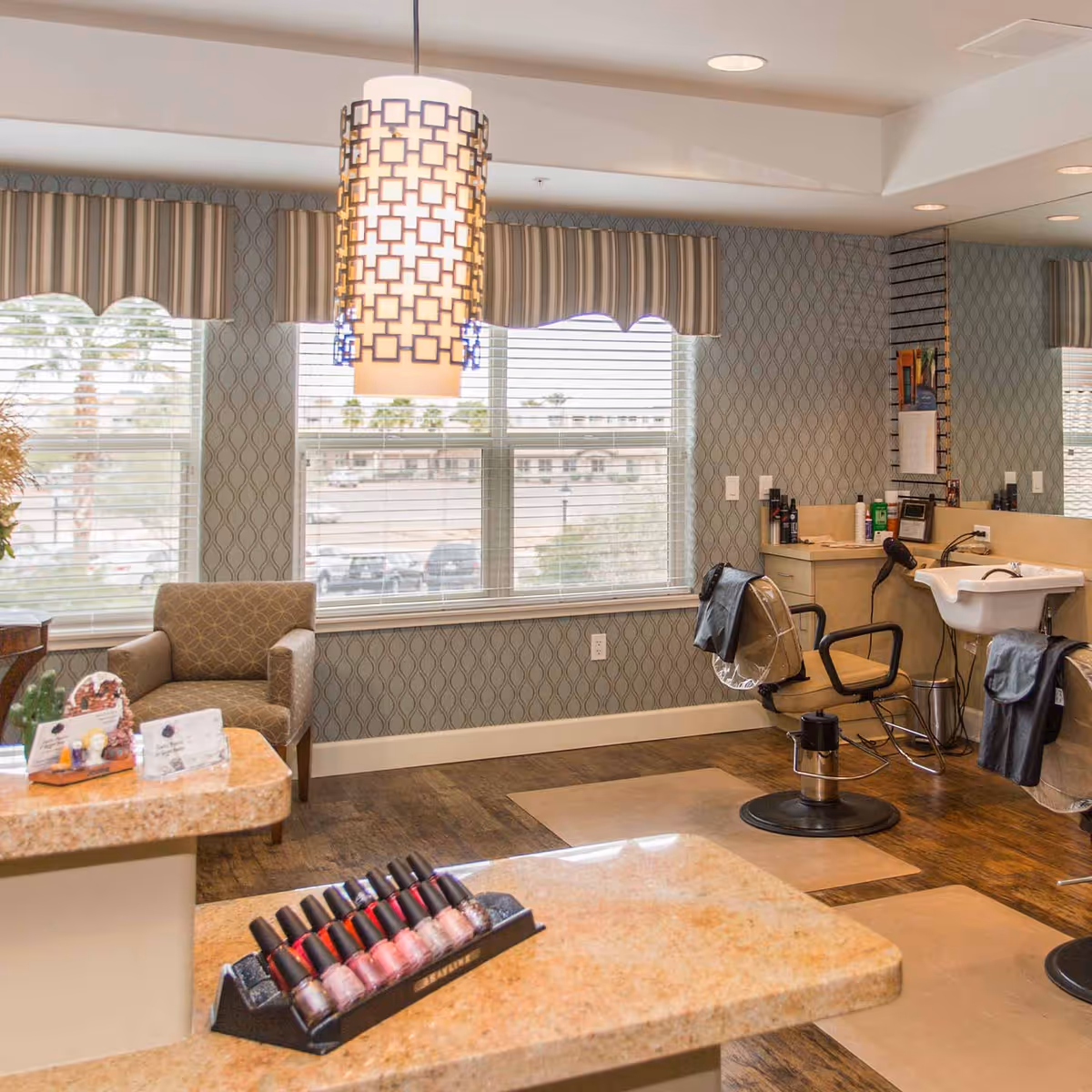 Interior of a salon area in an assisted living community featuring a styling chair with a hair washing sink, a comfortable armchair near large windows with striped valances, a hanging decorative light fixture, and a countertop displaying a selection of nail polish bottles.