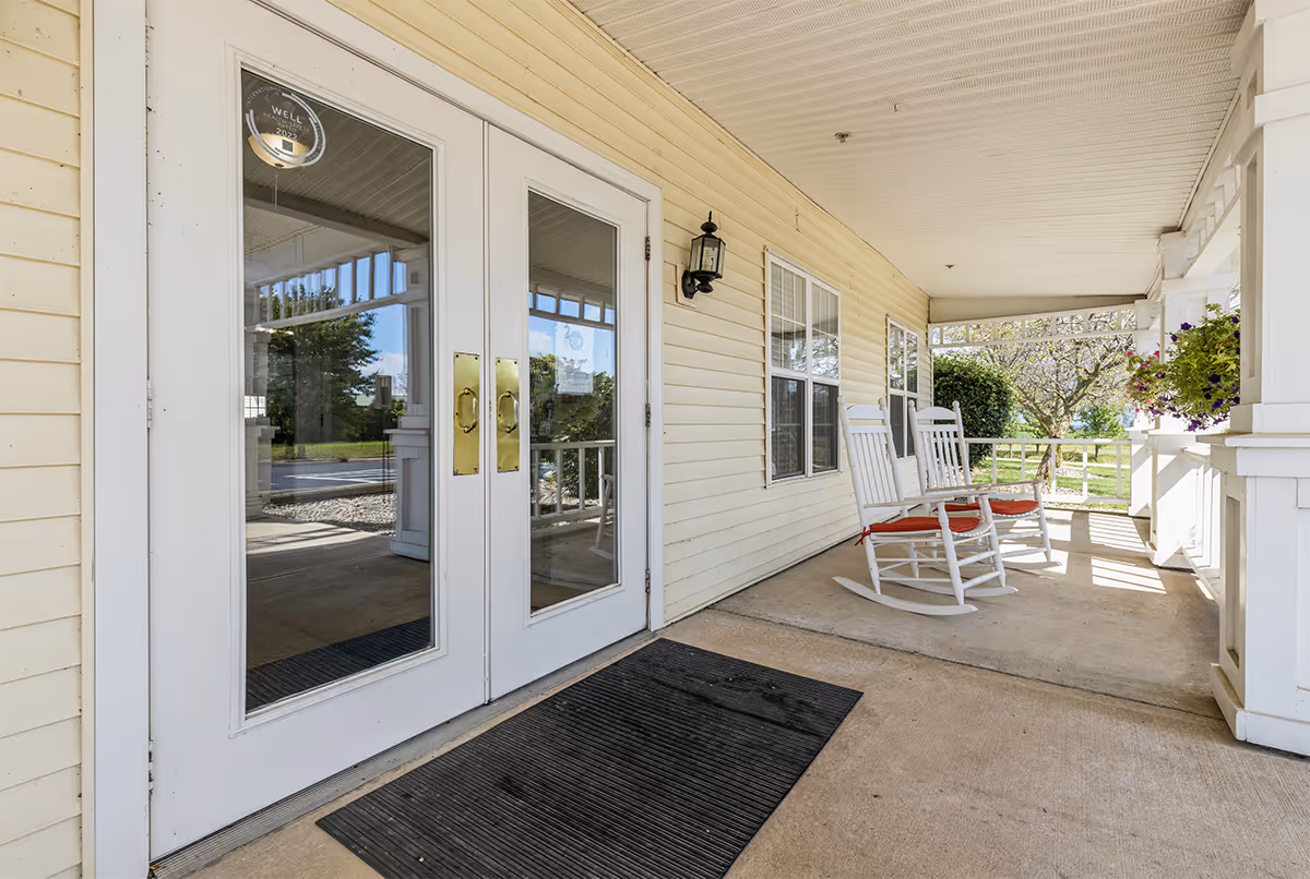 Covered front porch with double glass doors, two white rocking chairs with red cushions, and hanging flowers at the entrance.
