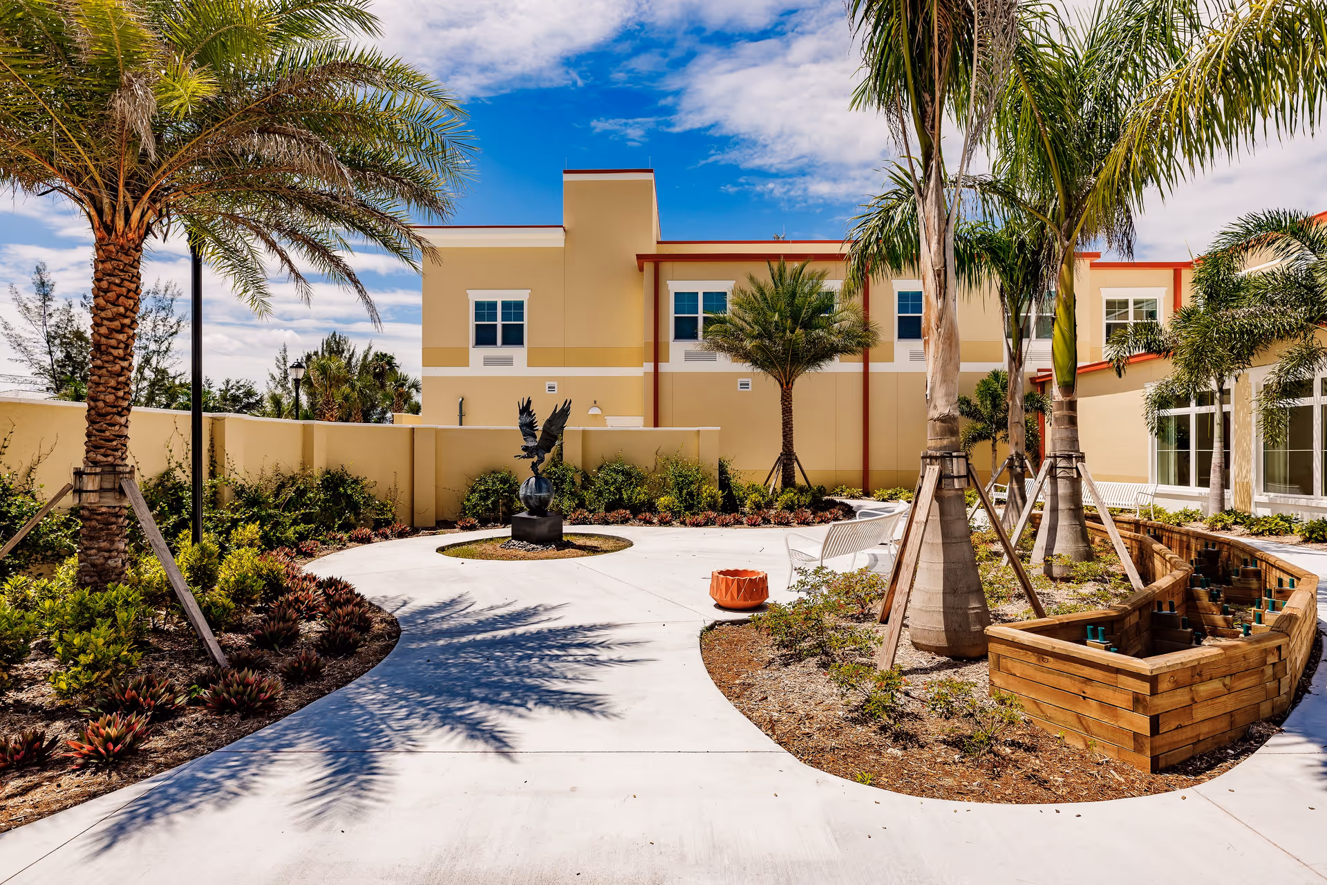 Outdoor courtyard area at The Gallery at Cape Coral featuring a curved concrete pathway, palm trees, landscaped garden beds, a metal eagle sculpture on a pedestal, white benches, and a wooden planter box under a partly cloudy blue sky.