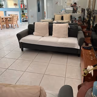 A living room area with a dark gray sofa covered partially with a white sheet and three beige cushions. Behind the sofa is a large mirror reflecting the room and a person taking the photo. To the left, there is a dining area with a wooden table and chairs near large windows letting in natural light. The floor is tiled with light-colored tiles.