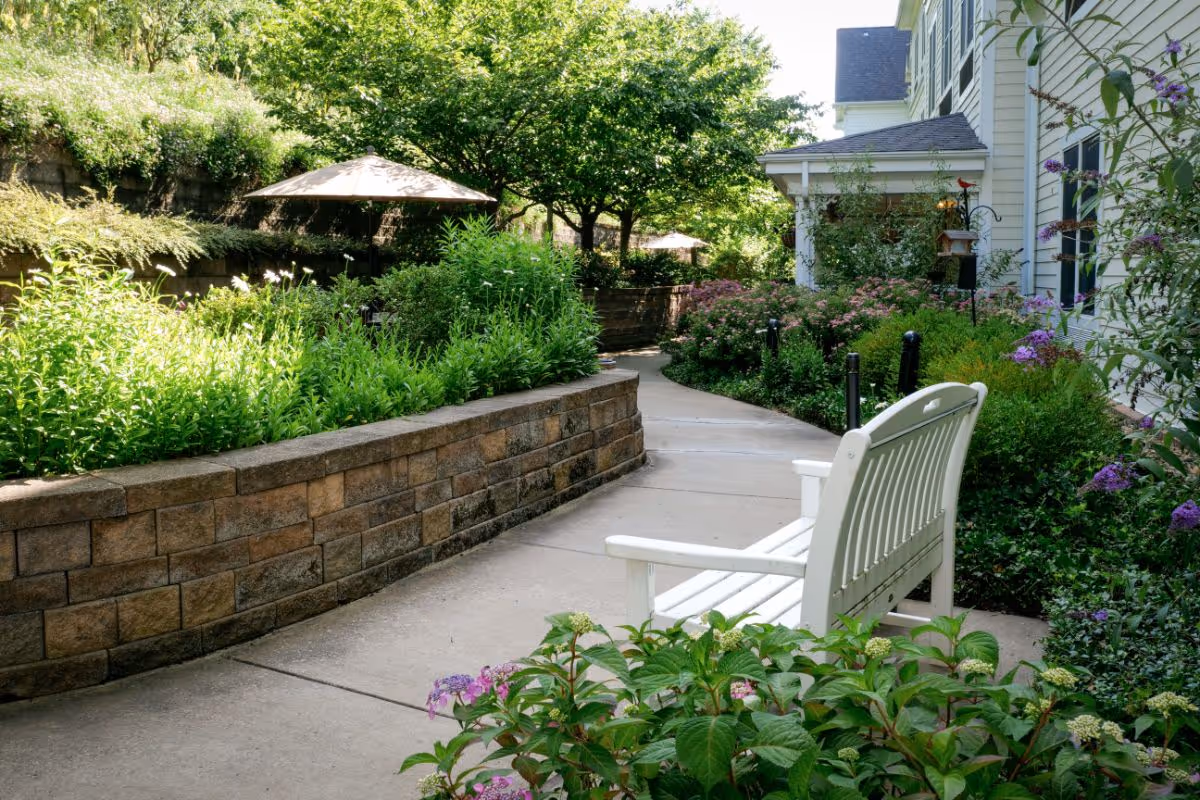 A peaceful outdoor garden area at Hemsley House of Upper St. Clair featuring a curved concrete pathway, a white bench, lush green plants, flowering bushes, and trees providing shade. There are umbrellas in the background and the side of a light-colored building is visible on the right.