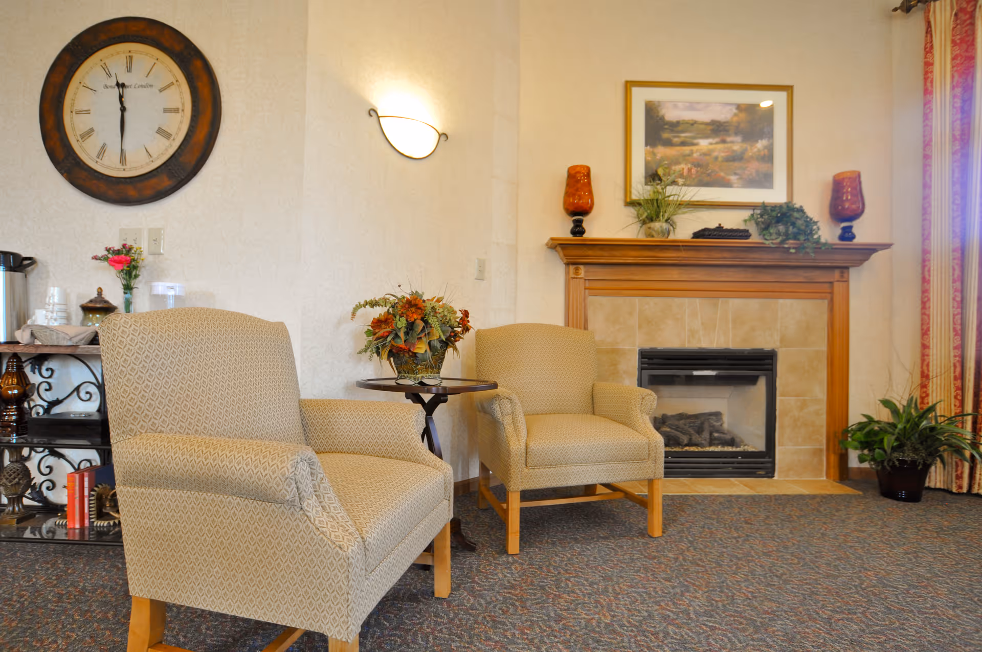 A cozy sitting area with two beige upholstered armchairs facing each other, a small round table with a floral arrangement between them, a fireplace with a wooden mantle decorated with plants and orange glass candle holders, a large round wall clock, a wall sconce light, and a small shelf with a coffee setup and decorative items.