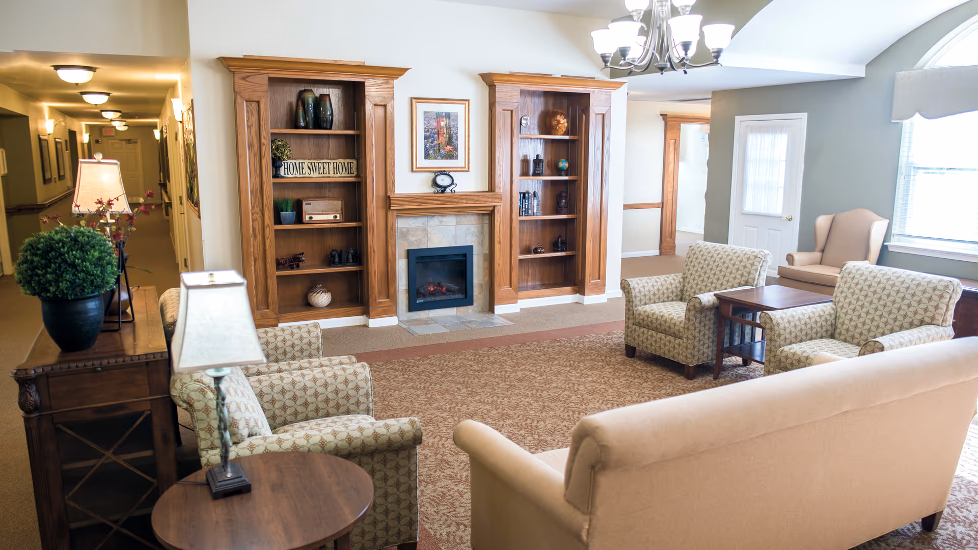 Bright senior living lounge with sofas and armchairs arranged around a fireplace and built-in wooden shelving.