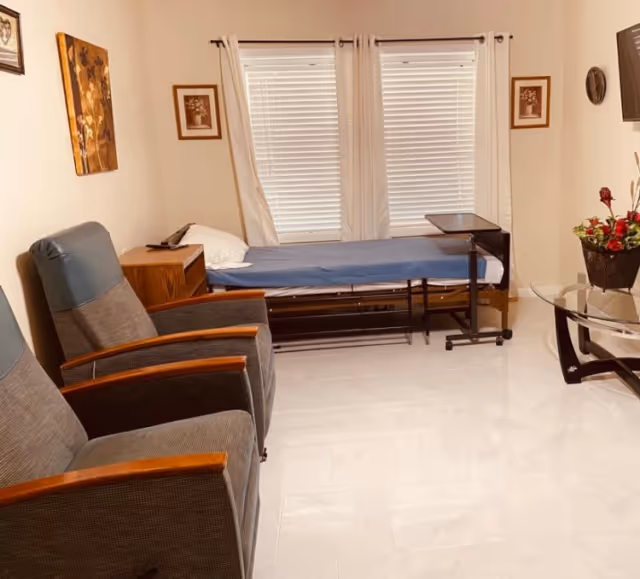 A small room with two gray armchairs with wooden armrests, a single bed with a blue cover and white pillow, a wooden nightstand, a small overbed table, two windows with white blinds and curtains, a glass coffee table with a flower arrangement, and framed artwork on the walls.