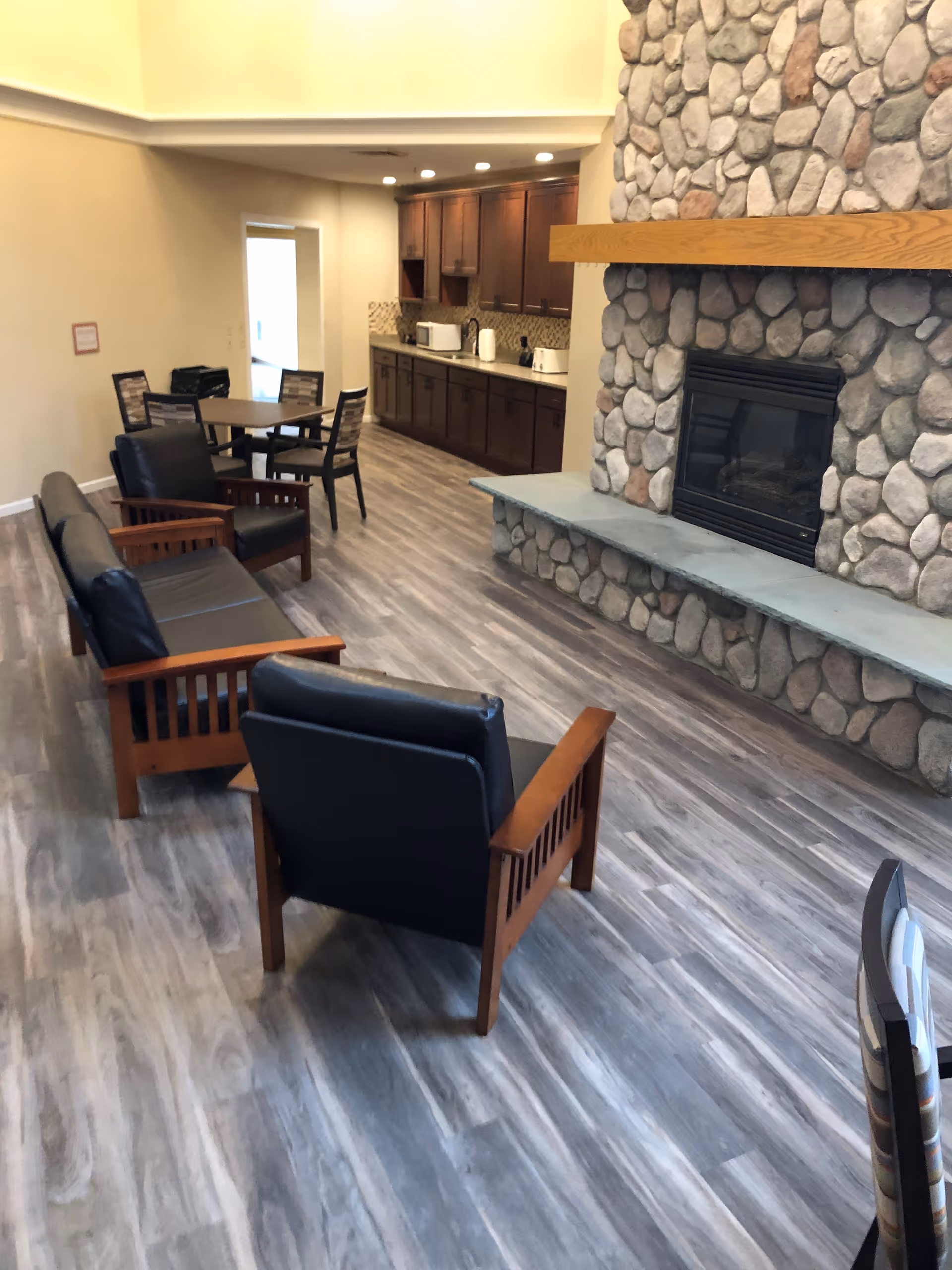 Interior view of a senior living facility common area featuring a stone fireplace with a wooden mantel, wooden framed chairs with black cushions, a wooden sofa with black cushions, a round table with chairs, and a kitchenette with dark wooden cabinets and appliances in the background.