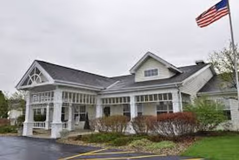 White Maplecrest Assisted Living building with porch, flagpole, and manicured shrubs.
